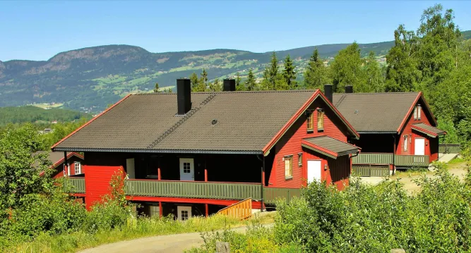 Ein großes rotes Holzhaus mit grauem Dach und Balkonen steht am Hang, umgeben von Bäumen vor Bergen..