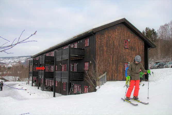 Ein Skifahrer mit Helm und Skistöcken steht auf einer verschneiten Piste vor einem großen dunklen Holzhaus..