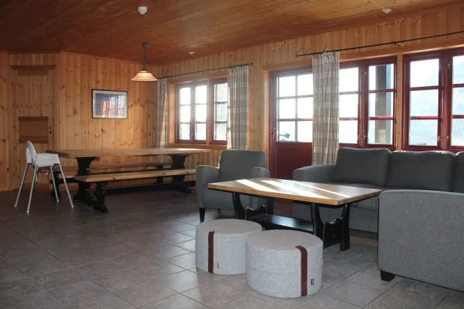 Holzvertäfelter Raum mit grauem Sofa, Sesseln, Couchtisch, zwei Hockern und Esstisch neben großen Fenstern..