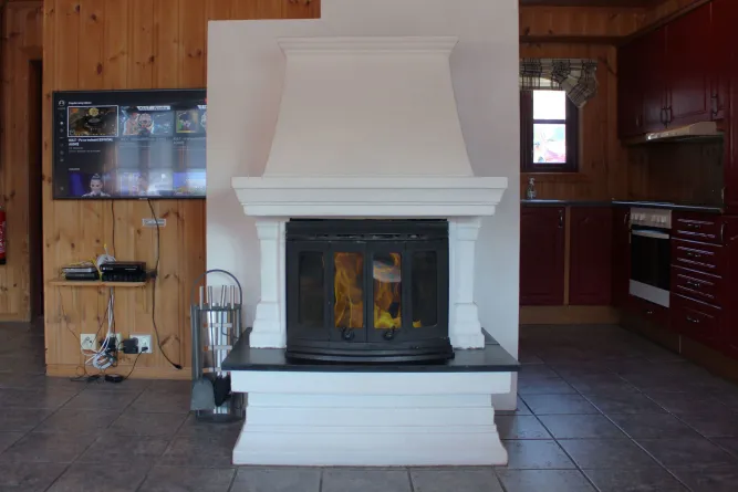 Weißer Kamin mit Glastür und Feuer, links ein Fernseher an Holzvertäfelung, rechts eine kleine Küche mit Fenster..