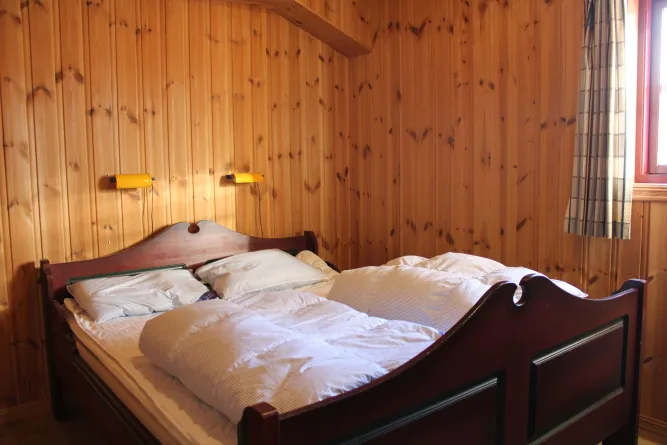 Holzvertäfeltes Schlafzimmer mit Doppelbett, weißen Bettdecken, zwei Wandlampen und Fenster mit kariertem Vorhang..