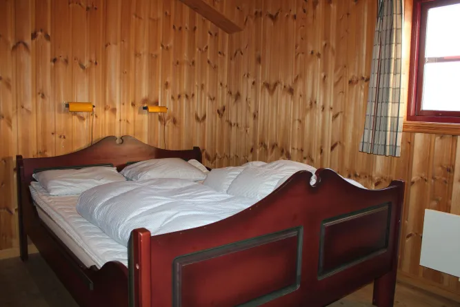 Holzvertäfeltes Schlafzimmer mit Doppelbett, weißer Bettwäsche, zwei Wandlampen und Fenster mit kariertem Vorhang..