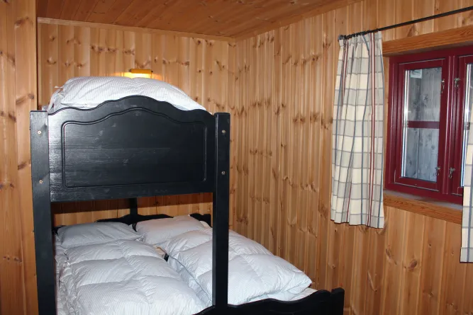 Holzvertäfeltes Schlafzimmer mit schwarzem Etagenbett, weißer Bettwäsche, kleinem Wandlicht und Fenster mit Vorhang..