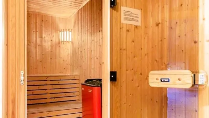 Innenansicht einer Holzsauna mit Sitzbank, Lampe an der Wand und rotem Saunaofen neben der Tür..
