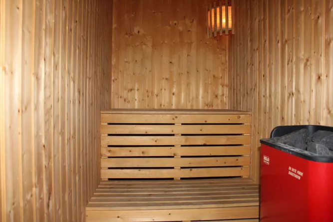 Innenansicht einer Holzsauna mit Sitzbank, Wandlampe und rotem Saunaofen mit Saunasteinen rechts..