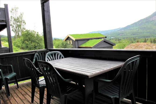 Schwarzer Terrassentisch mit Plastikstühlen unter Überdachung, dahinter Hütte mit Grasdach und Berge..