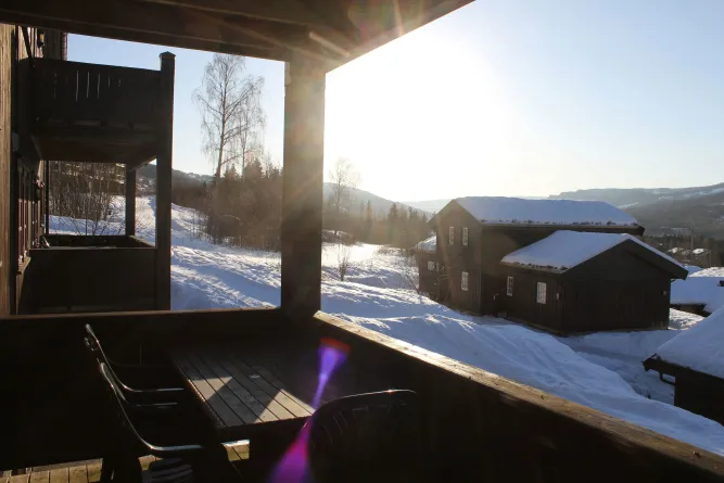 Blick von einem Holzbalkon mit Tisch und Stühlen auf verschneite Hütten, Bäume und Hügel im Gegenlicht..
