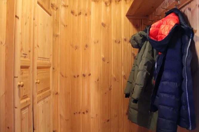 Holzvertäfelter Flur mit Schranktüren links und mehreren Winterjacken, die rechts an Haken unter einem Regal hängen..