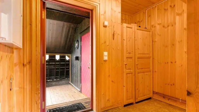 Holzvertäfelter Eingangsbereich mit Kleiderschrank, offener Tür und Blick in einen überdachten Vorraum mit roter Tür..