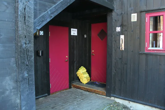 Überdachter Holzhaus-Eingang mit zwei roten Türen, schwarzer Fassade, rotem Fenster und einem Sack Brennholz auf der Stufe..