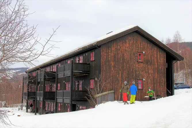 Dunkles Holzhaus mit roten Fenstern im Schnee; davor stehen drei Skifahrer in bunter Winterkleidung am Hang..