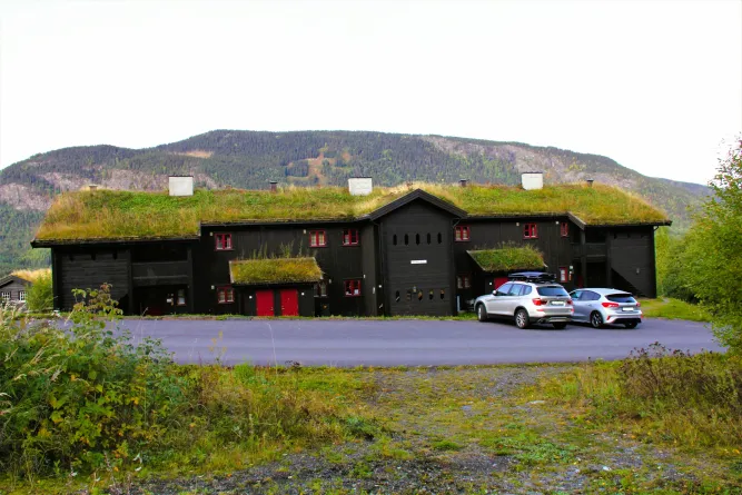 Schwarzes Holzgebäude mit Grasdach und roten Fenstern, davor zwei geparkte Autos, im Hintergrund Berge..