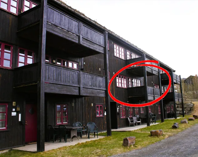 Dunkles Holzgebäude mit roten Fensterrahmen und Balkonen; ein Bereich rechts ist mit rotem Kreis markiert..