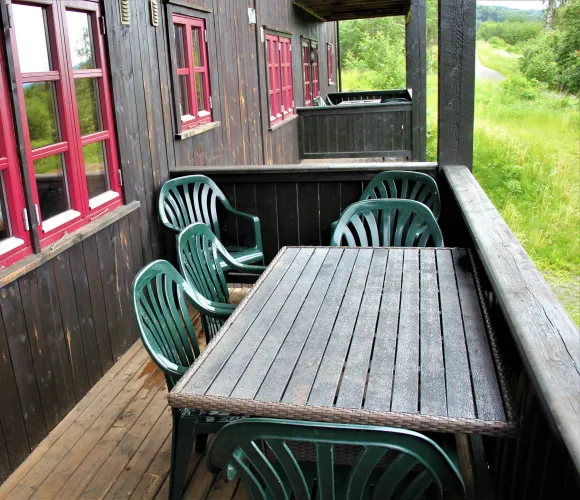 Holzterrasse an dunkler Hütte mit rotem Fensterrahmen, Tisch und grünen Kunststoffstühlen neben Wiese..