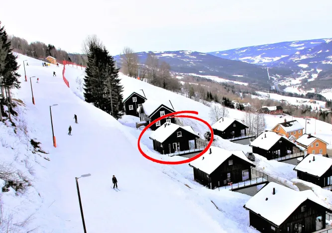 Besneeuwde skipiste langs zwarte houten chalets; een huis is rood omcirkeld, met skiërs en bergen op de achtergrond..