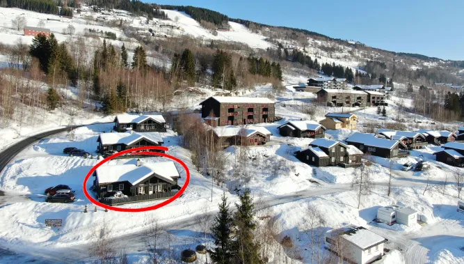 Luchtfoto van een besneeuwd vakantiedorp met houten chalets en wegen; één chalet is rood omcirkeld..