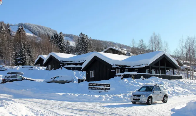 Donkere houten chalets met dikke sneeuw op het dak langs een besneeuwde weg, auto op de voorgrond en bergen erachter..