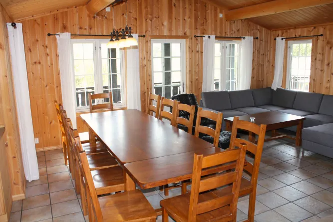Houten interieur met lange eettafel en stoelen, hoekbank en salontafel bij ramen met witte gordijnen..