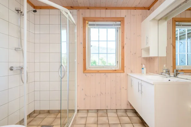 Heldere badkamer met glazen douchecabine, raam met houten lijst en wit wastafelmeubel met spiegel..