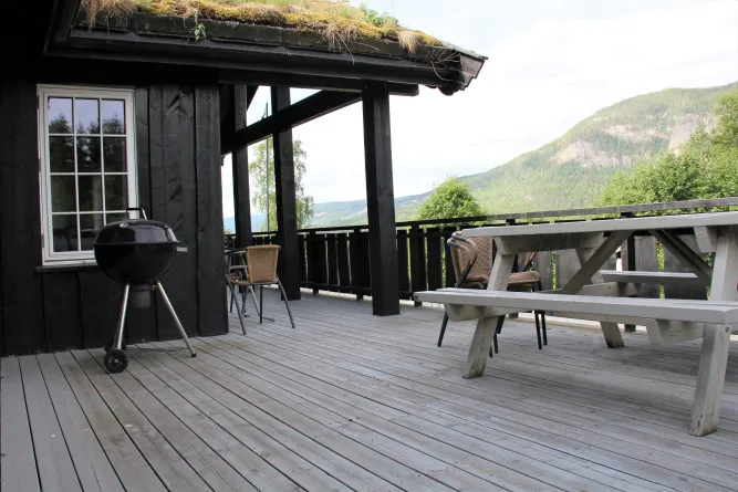 Houten veranda bij zwart huis met barbecue, stoelen en picknicktafel, met berglandschap op de achtergrond..