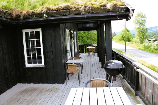Overdekte houten veranda met tafels, rieten stoelen en een ronde barbecue naast een zwarte hut met grasdak..