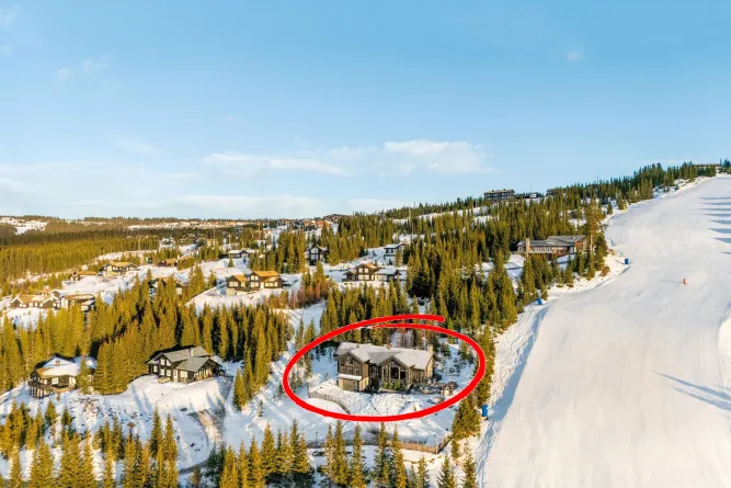 Luchtfoto van een besneeuwd bergdorp met dennenbos en chalets; een huis is rood omcirkeld naast een skipiste..