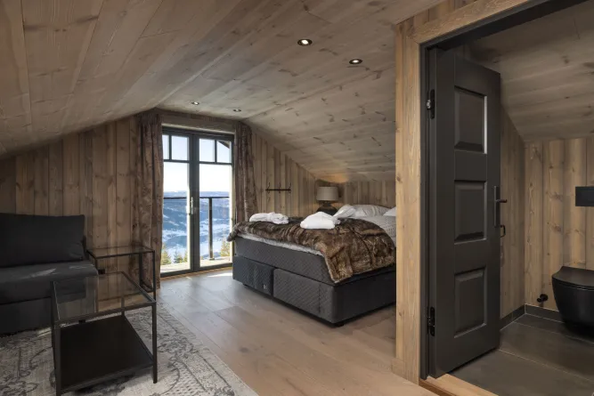 Gezellige houten zolderslaapkamer met tweepersoonsbed, bank en grote ramen met uitzicht op een besneeuwd landschap..