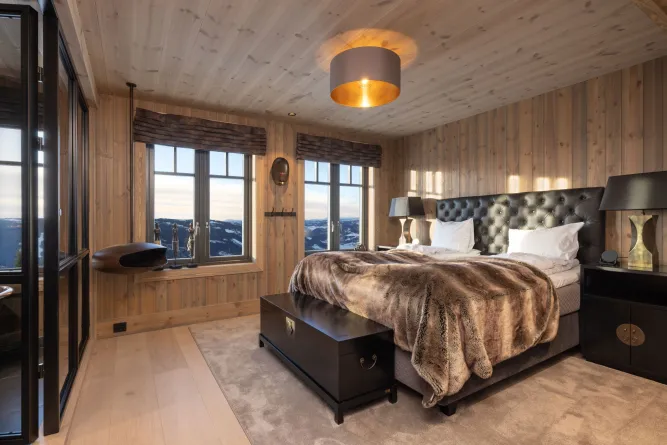 Houten slaapkamer met tweepersoonsbed, zwarte nachtkastjes en grote ramen met uitzicht op besneeuwde bergen..