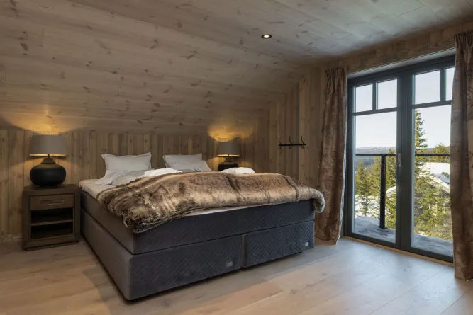 Houten slaapkamer met tweepersoonsbed, nachtkastjes en lampen, naast een groot raam met uitzicht op besneeuwde bomen..