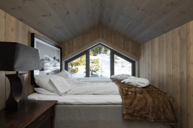 Houten slaapkamer met tweepersoonsbed, lamp en groot raam; buiten zijn besneeuwde bomen en helling zichtbaar..