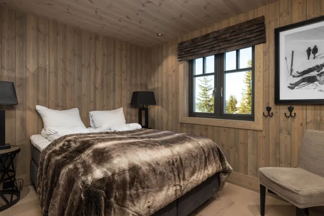 Houten slaapkamer met tweepersoonsbed en bontdeken, zwarte lampen, raam met uitzicht op bomen en een stoel..