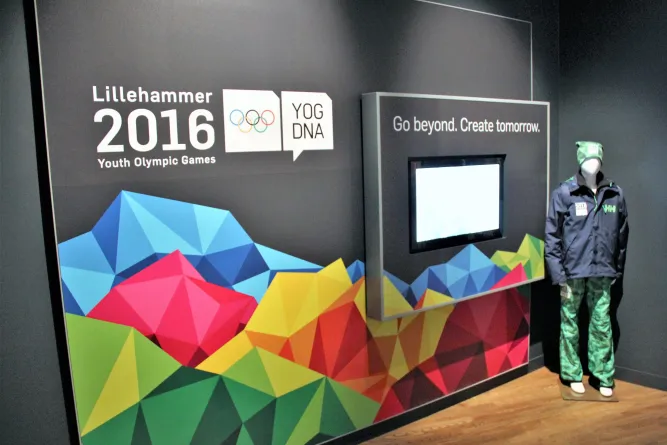 Ausstellungswand zu „Lillehammer 2016 Youth Olympic Games“ mit buntem Bergmotiv, Bildschirm und Schaufensterpuppe..