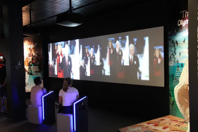 Zwei Personen mit Kopfhörern sitzen in einem Ausstellungsraum und sehen eine breite Videoprojektion an der Wand..