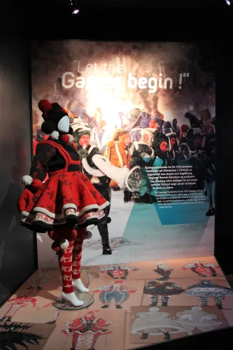 Puppe in rotem Kostüm steht in einer Ausstellung vor einem Foto einer jubelnden Wintermenge mit Schriftzug..