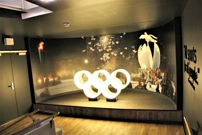 Beleuchtete olympische Ringe auf Podest vor großem Wandbild; dunkler Museumsraum mit Türen und Schriftzug rechts..