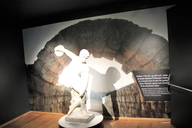 Beleuchtete weiße Statue in einem Innenraum vor einem großen Steinbogen mit Infotafel an der rechten Wand..