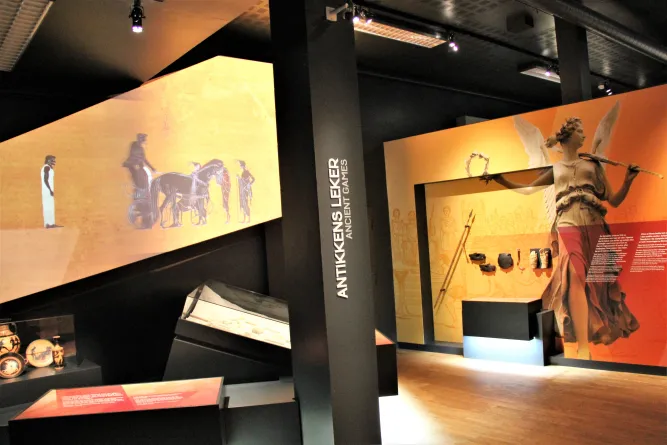 Innenansicht einer Ausstellung mit antiken Wandbildern, Vitrinen und einer großen Engelsfigur vor orangefarbenem Hintergrund..