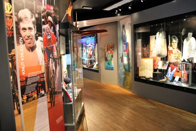 Innenansicht eines Museums mit Holzfußboden, Sportpostern, Glasvitrinen und ausgestellten Sportobjekten an den Wänden..