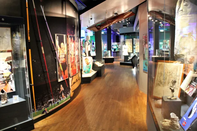 Beleuchteter Museumsflur mit Vitrinen, Plakaten und Sportexponaten; an der Decke hängt ein schmales Boot..