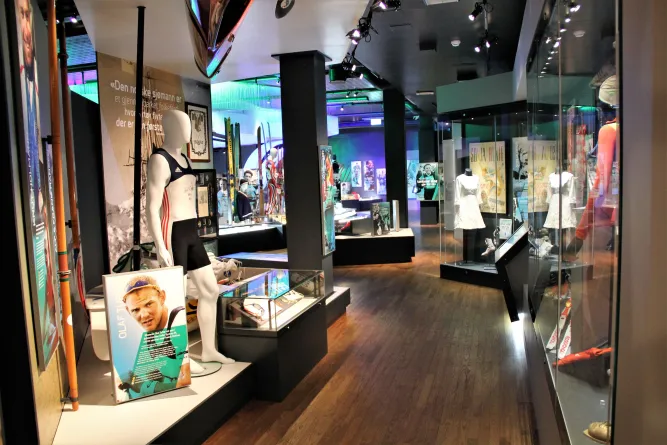 Blick in eine Museumsausstellung mit Schaufenstern, Skiern, Sportkleidung auf Schaufensterpuppen und Beleuchtung an der Decke..