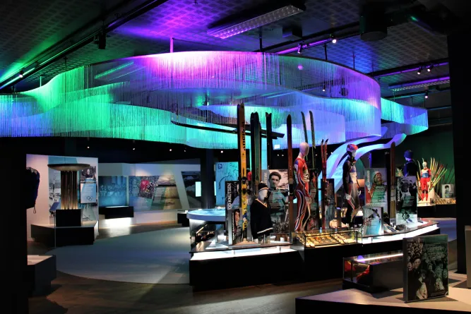 Innenraum einer Ausstellung mit farbiger Deckeninstallation, Vitrinen, Skiern und Schaufensterpuppen in Sportkleidung..