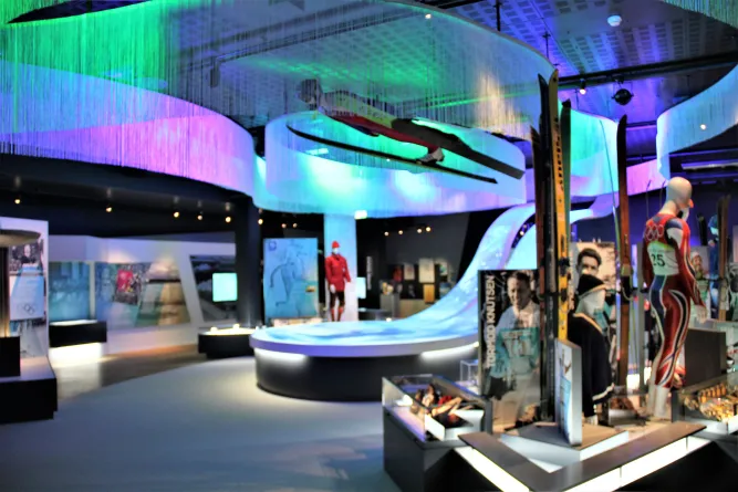Innenansicht eines Museums mit farbiger Beleuchtung, ausgestellten Skiern, Vitrinen und Schaufensterpuppen in Sportkleidung..