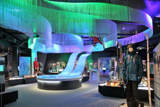 Museumsraum mit farbig beleuchteter Deckeninstallation, Ski-Exponaten, Vitrinen und einer bekleideten Schaufensterpuppe..