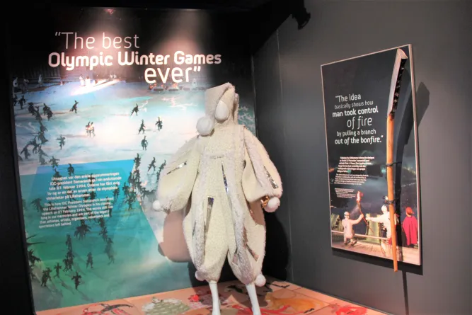 Ausstellungsraum mit Wandzitaten zu Olympischen Winterspielen und einer weißen, kapuzenartigen Kostümfigur..