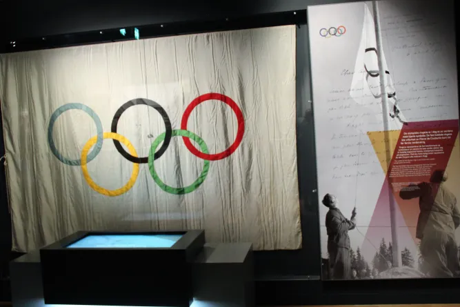 Ein großer Stoff mit den olympischen Ringen hängt an der Wand; daneben steht eine Infotafel in einem Museum..