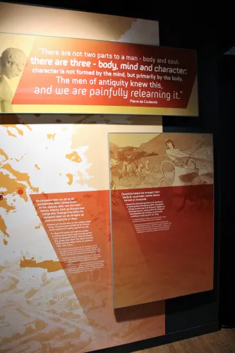 Wanddisplay in einem Museum mit großem englischen Zitatbanner, Karte und Infotafeln zu den antiken Olympischen Spielen..