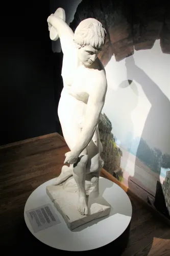 Weiße Skulptur eines nackten Jünglings auf rundem Podest im Innenraum, stark beleuchtet mit deutlichem Schatten..