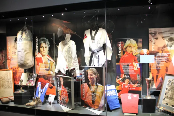 Museumsvitrine mit Sportexponaten wie Boxsack, Fechtmaske, Kampfsportanzug, Medaillen und Handballplakat..