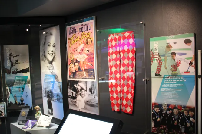 Museumsausstellung mit Sportplakaten, Info-Tafeln und rot-weiß gemusterter Hose hinter Glas an dunkler Wand..
