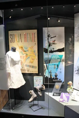 Museumsvitrine mit weißem Eiskunstlaufkleid, Schlittschuhen, Plakaten und Ausstellungsstücken hinter Glas..